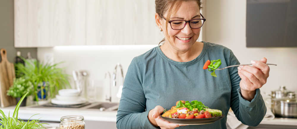 Middle-aged woman smiling, about to take a bite of a fresh salad in her kitchen