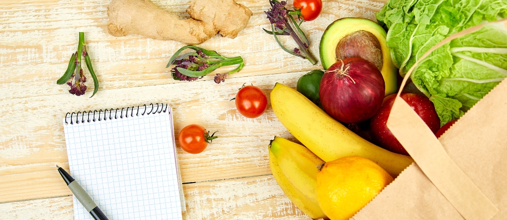 Top view of notepad and pen for writing a shopping list and grocery bag full of fresh produce