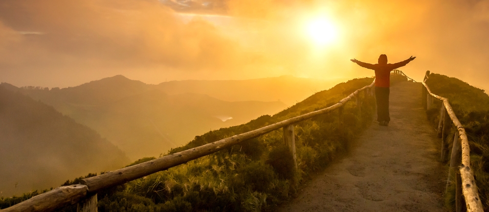 Person with arms raised in victory, standing along a path atop a mountain with the sun shining brightly.
