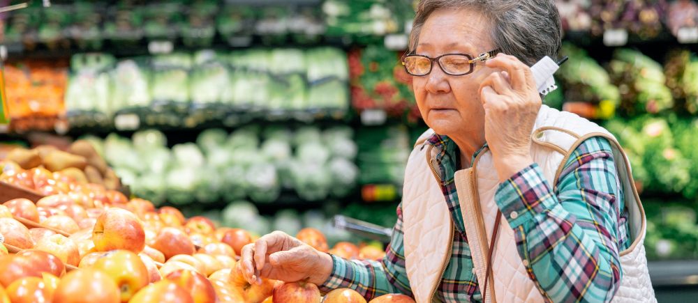 Older adult woman raising glasses to examine produce in a grocery store.
