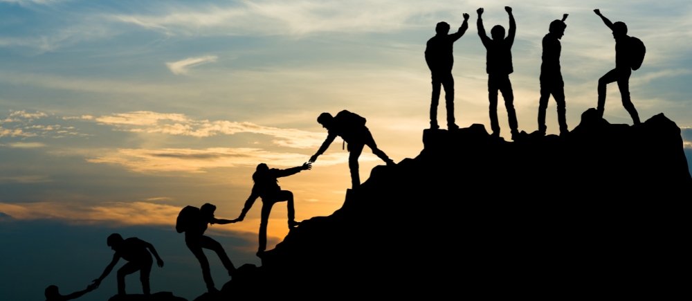 A silhouette of a group of hikers at sunset, extending helping hands to assist each other in reaching the mountain's summit.