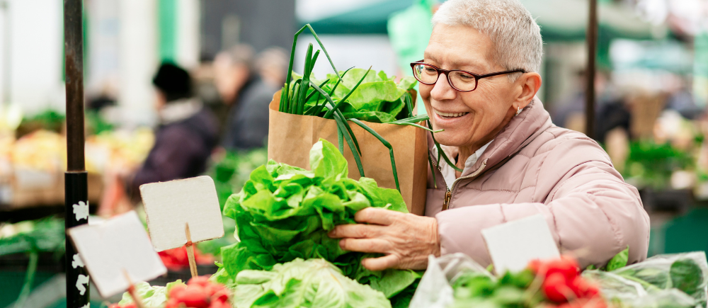 An older woman smiling as she holds a bunch of fresh lettuce and a brown paper bag filled with produce at an outdoor market.